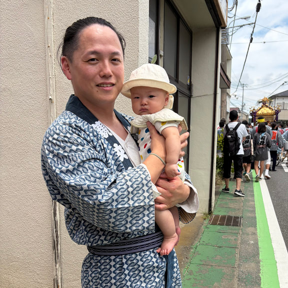 木更津の祭りで、子どもを抱いて神輿の列のそばに立つ代表の東